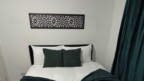 Ein ordentlich bezogenes Bett mit weißer und grüner Bettwäsche, ein dekoratives Wandbild darüber und ein dunkelgrüner Vorhang zur Rechten.
