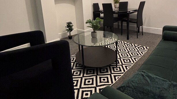 Ein modernes Wohnzimmer mit einem grünen Sofa, schwarzen Sesseln, einem Glastisch, einem Esstisch mit Stühlen, einem flachbildfernseher an der Wand und einem gemusterten Teppich.