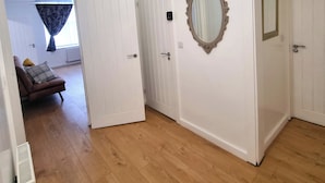 A hallway with white walls, wooden flooring, and a door leading to a room with a window and curtains. - Forge Lane Cozy Apartment (Gillingham)