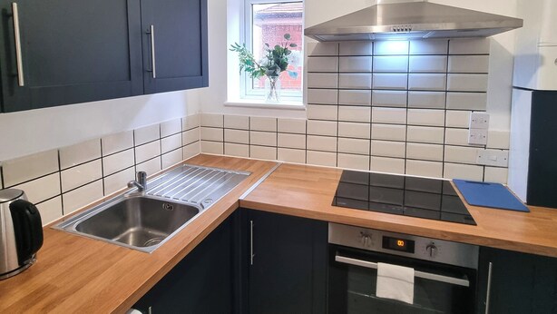 A modern kitchen with dark cabinets, a stainless steel sink, a built-in oven, and a window with a view of the outside. - Forge Lane Cozy Apartment (Gillingham)