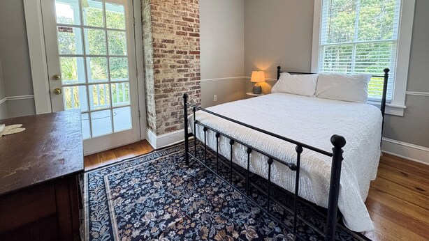 A bedroom with a brick accent wall, a bed with white bedding, a nightstand with a lamp, and a large window with blinds. - Historic Beaufort Estate - Near Downtown Beaufort & Parris Island (Beaufort)