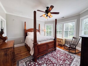 A four-poster bed with a wooden headboard, a ceiling fan, and a red floral area rug.