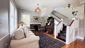 A spacious living room with a sofa, a fireplace, a staircase, and a chandelier. - Historic Beaufort Estate - Near Downtown Beaufort & Parris Island (Beaufort)