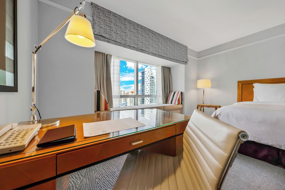 A hotel room with a desk, a bed, a window with curtains, and a view of buildings.
