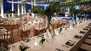 Un espace extérieur aménagé pour les repas, avec des guirlandes lumineuses, des palmiers et des tables dressées pour un événement formel.