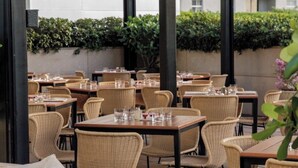 Un espace de restauration en plein air avec des chaises et des tables en osier, des lampes en osier suspendues au-dessus, et une vue sur un bâtiment en arrière-plan.