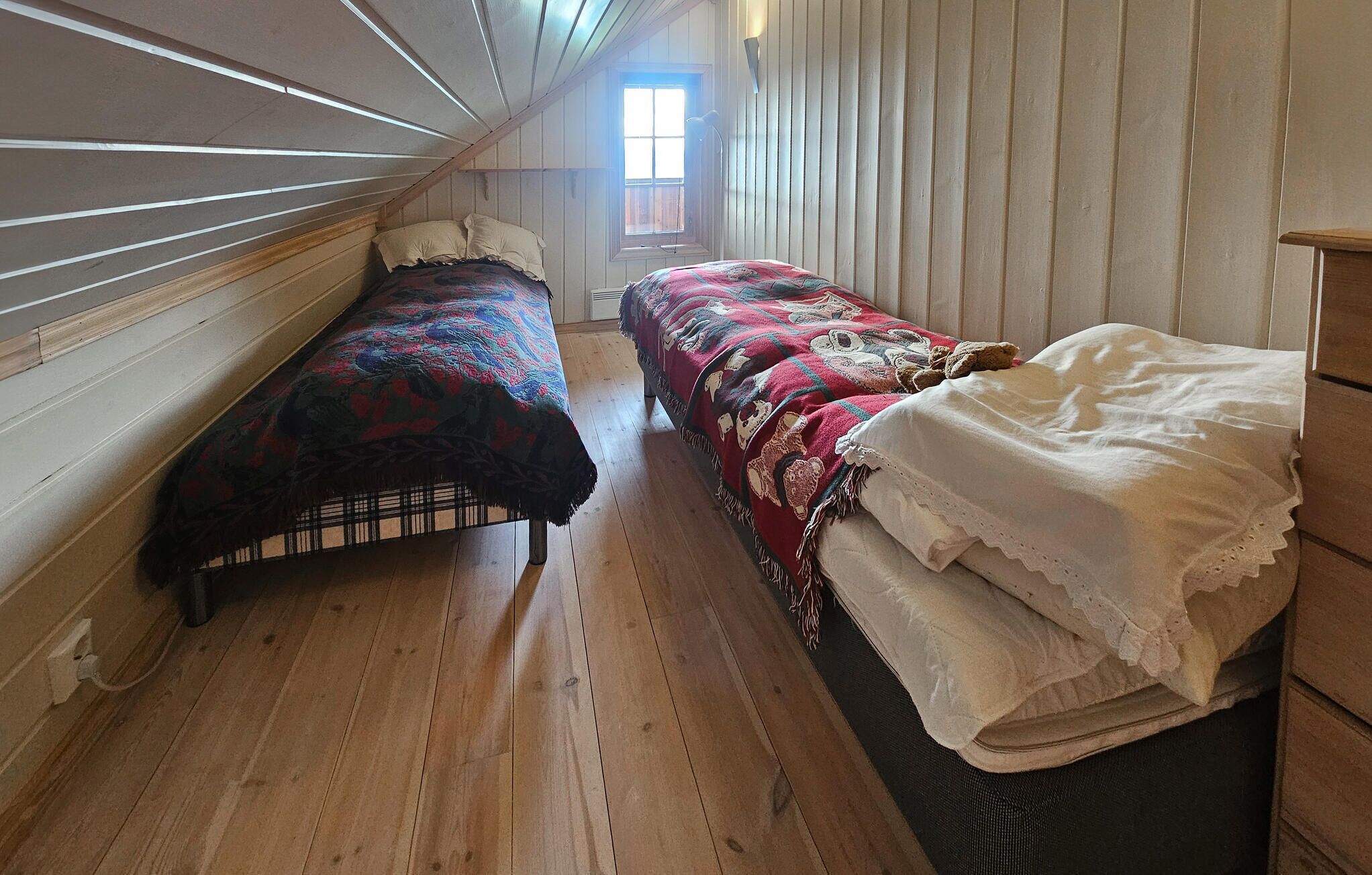 Ett smalt rum med två sängar, trägolv och ett fönster som släpper in naturligt ljus.