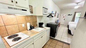 A compact kitchen with a stove, sink, and microwave. There is a small refrigerator, a coffee maker, and a black cabinet. The room has a tiled floor and white walls.