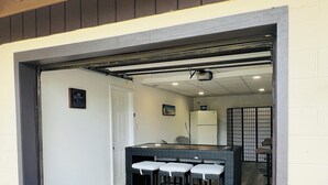 A covered outdoor seating area with a table and bar stools, a refrigerator, and a door with a sign.