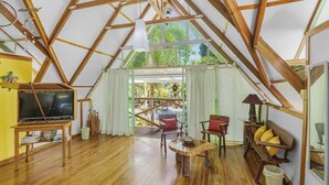 Una habitación amplia con techo alto, vigas de madera, un televisor sobre un mueble bajo, una mesa de comedor y vistas a zonas verdes.