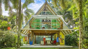 Una casa triangular con fachada de vidrio y terraza de madera, rodeada de abundante vegetación.