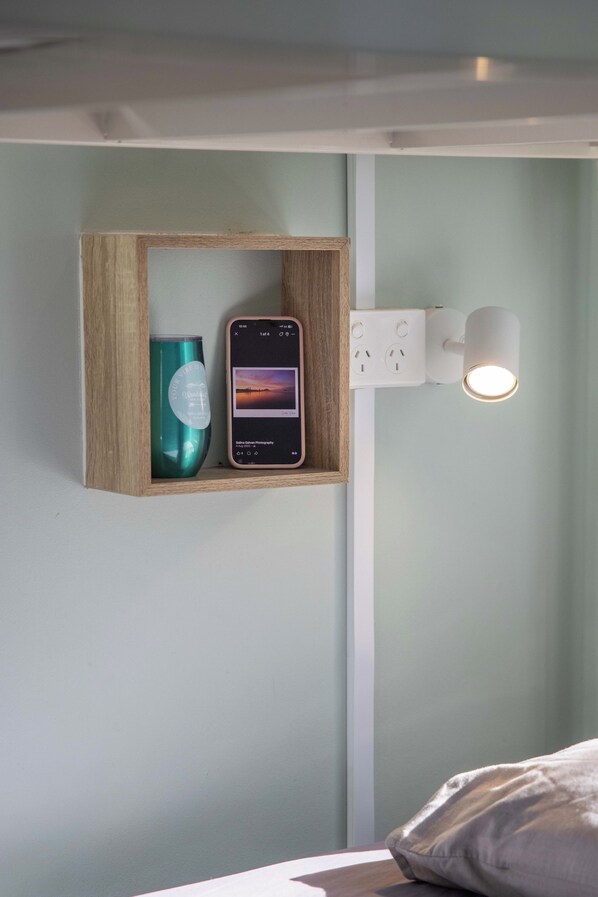 A smartphone mounted on a wall with a wooden shelf below it, a wall socket, and a light fixture. - Wanderlust NZ (Tauranga)