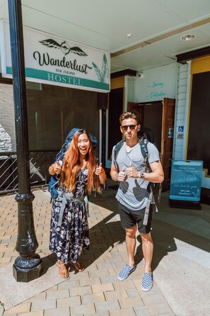 Two people standing in front of a hostel named 'Wanderlust New Zealand Hostel' with a signboard indicating availability for dorms, twins, and doubles with ensuite. - Wanderlust NZ (Tauranga)