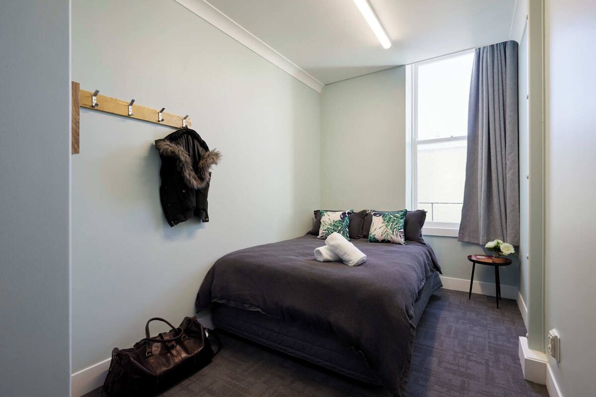 A bedroom with a bed, a coat rack with a fur-trimmed jacket, a small table with a vase, and a window with curtains.
