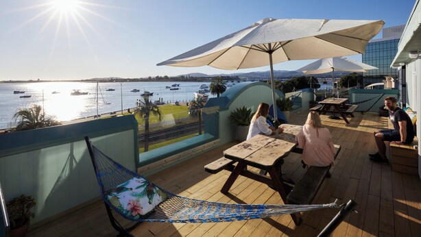 Una terraza con mesas y bancos de madera, una hamaca y sombrillas que dan a un cuerpo de agua con barcos.