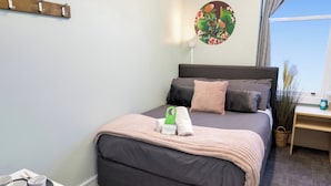 A neatly made bed with a grey headboard, a bedside table with a lamp, and a window with curtains. - Wanderlust NZ (Tauranga)