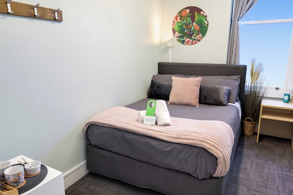 A neatly made bed with a grey headboard, a bedside table with a lamp, and a window with curtains. - Wanderlust NZ (Tauranga)