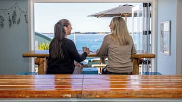 Two individuals seated at a wooden table with a view of the sea and boats in the background.