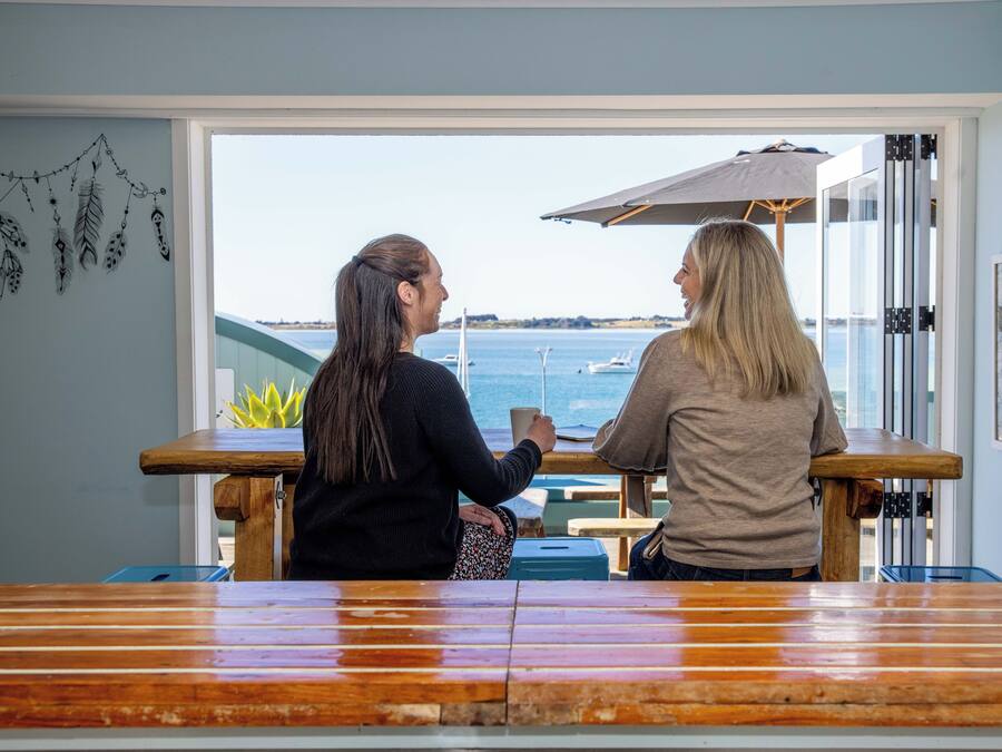 Zwei Personen sitzen an einem Holztisch mit Blick auf das Meer und Boote im Hintergrund.