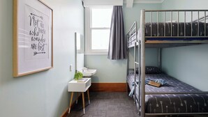 Una habitación pequeña y bien iluminada con literas, una mesita de noche con una planta, un espejo y una cita inspiradora enmarcada en la pared.