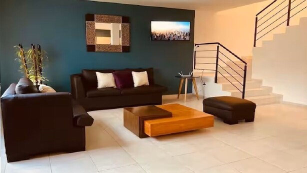 Ein modernes Wohnzimmer mit einem dunkelgrauen Sofa, einem Holztisch und einem kleinen Beistelltisch. Im Hintergrund ist eine Treppe zu sehen.