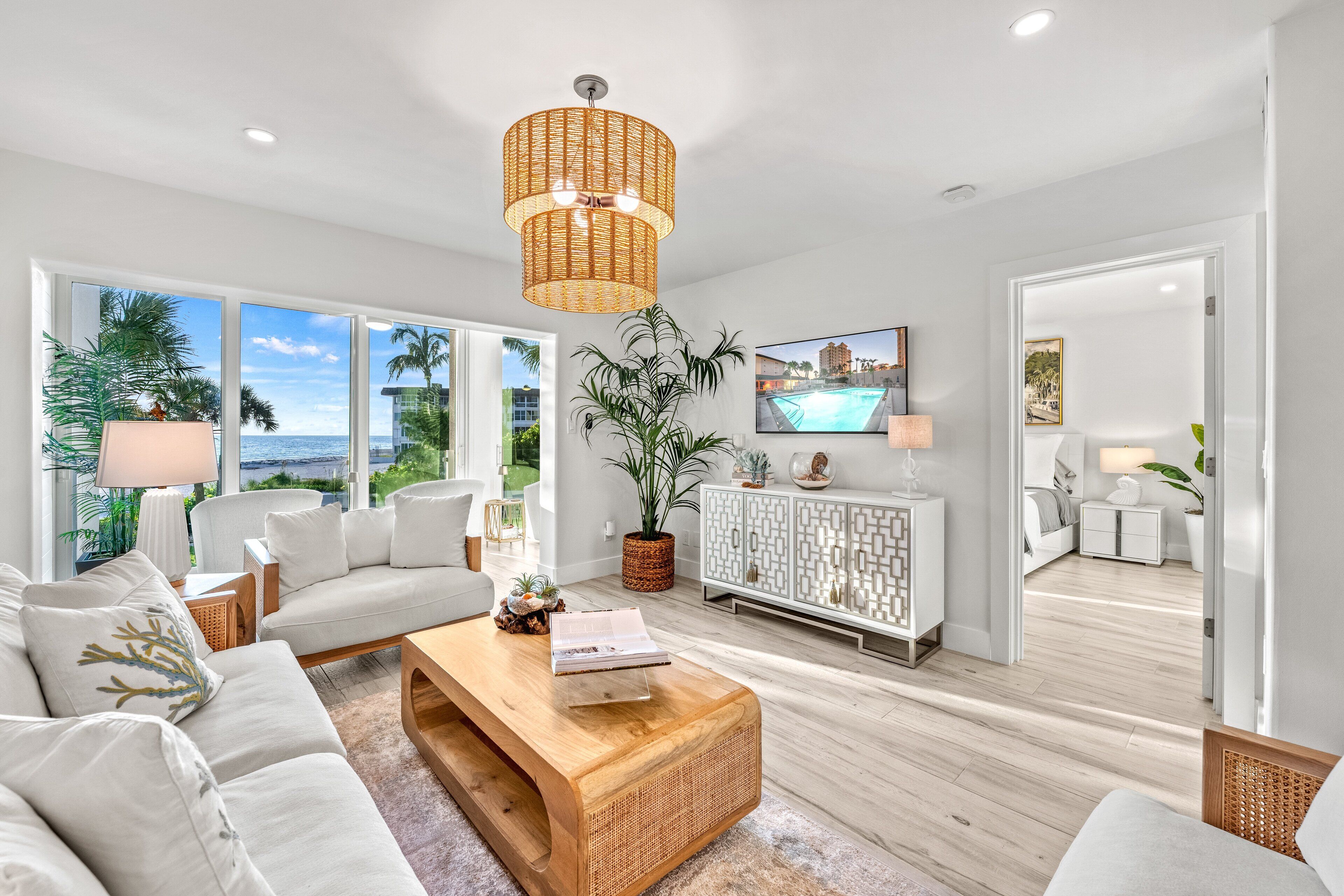A modern living room with a large window, a wicker coffee table, a white sofa, and a television on the wall.