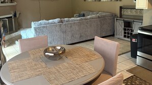 A modern living room with a grey sectional sofa, a round dining table with placemats, wicker chairs, a chandelier, a flat-screen TV, and a kitchen area with cabinets and a stove. - This cozy home is a beautiful 3 bedroom, 2 full bathroom as  with a 2-car garage (Baton Rouge)