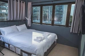 A neatly made bed with white linens and towels, a window with curtains, and a view of trees outside.