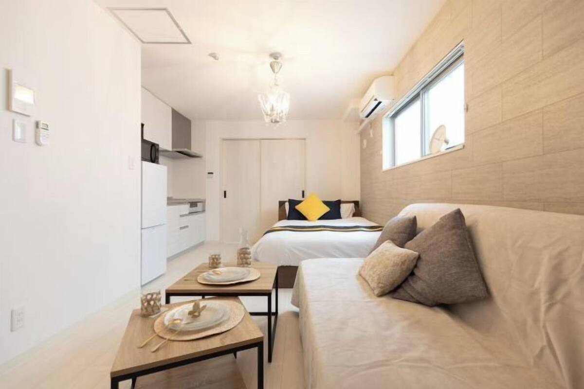 A modern hotel room with a bed, a sofa, a dining table with plates and cutlery, and a window with blinds.