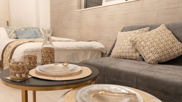 A modern living room with a grey sofa, patterned cushions, a round coffee table with a vase and plates, and a bed with a blue pillow. - Tengacyaya Ra Hotel (Osaka)