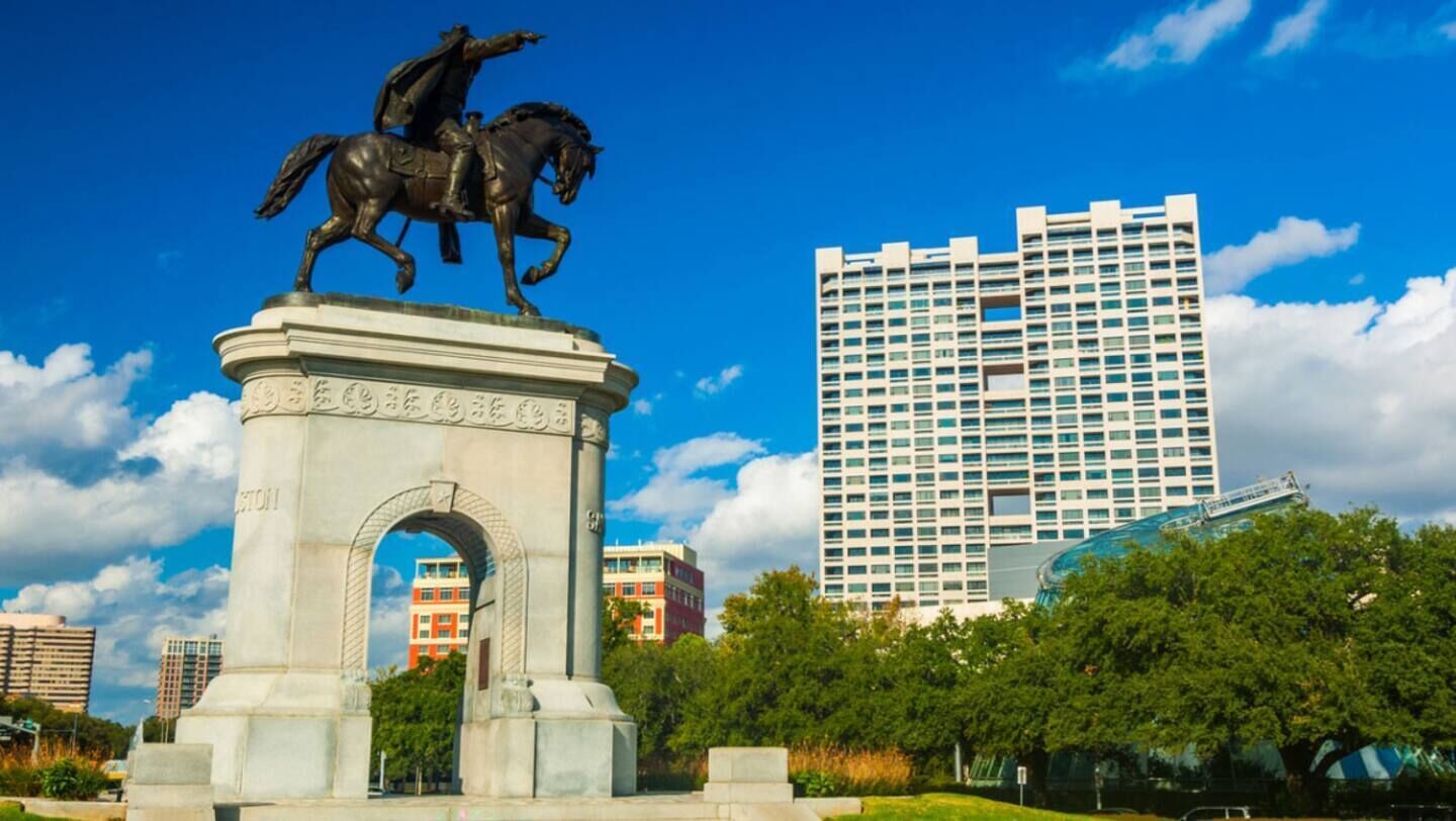 A statue of a figure on horseback atop a classical monument with an arch, surrounded by greenery and modern buildings in the background.