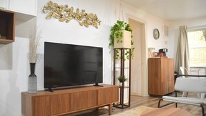 A living room with a large flat-screen TV on a wooden TV stand, a small round table with a remote, a tall plant in a white planter, and a wooden cabinet.
