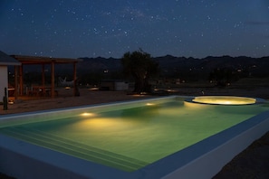 Una piscina al anochecer bajo un cielo estrellado, una estructura de madera y montañas al fondo.