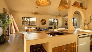 A spacious kitchen with a large island, hanging woven light fixtures, and a view through an archway to a living area. - Sundara | Pool | Spa | Pickleball | Sauna | Games (Joshua Tree)