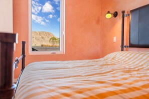 A bedroom with a bed, a window with a view of a desert landscape, a nightstand, and a wall-mounted lamp.
