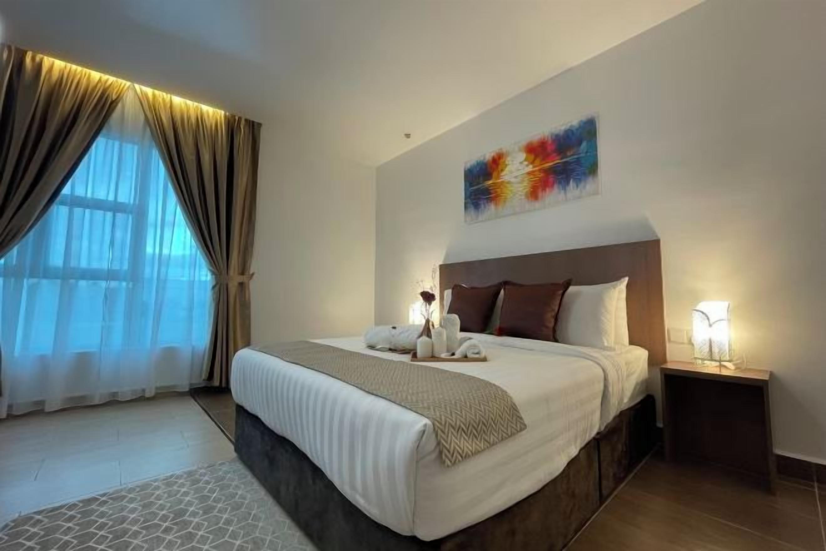 Ein Hotelzimmer mit einem großen Bett, einem Nachttisch, einem Wandbild und einem Fenster mit Vorhängen.