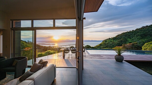 Ein modernes Interieur mit einer großen Glastür, die zu einem Balkon mit Blick auf den Sonnenuntergang über Wasser und Hügeln führt.