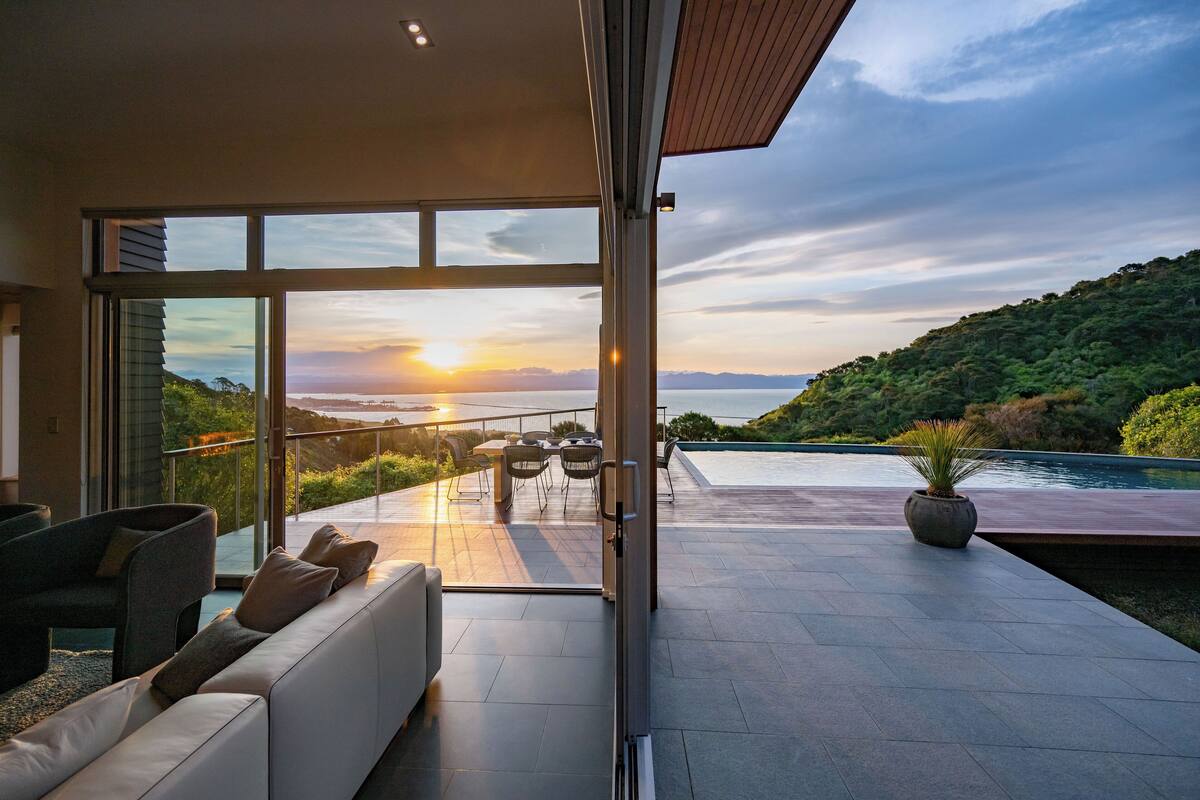 Un intérieur moderne avec une grande porte vitrée donnant sur un balcon d’où l’on peut admirer le coucher de soleil sur une étendue d’eau et des collines.