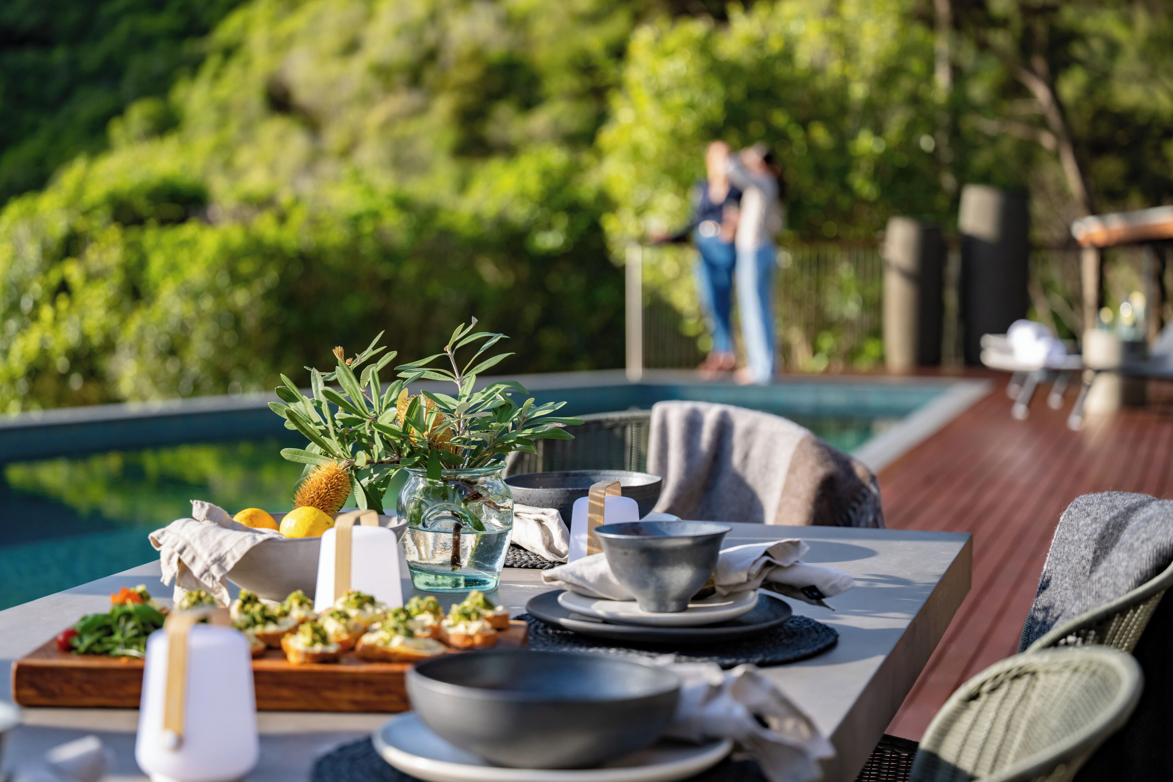 A dining table with a variety of food and drink items, set outdoors near a pool with a view of lush greenery and a person in the background.