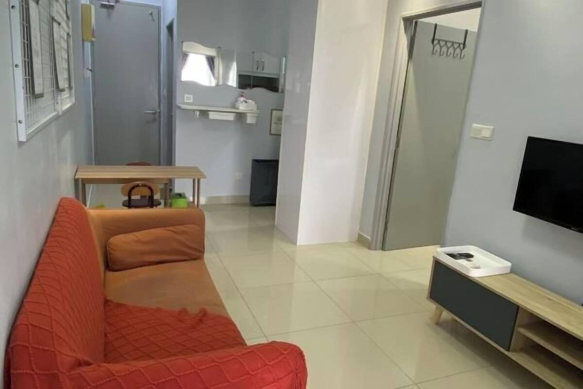 A modern living room with a red sofa, a small table with a kettle, a TV on the wall, and a kitchen area in the background.