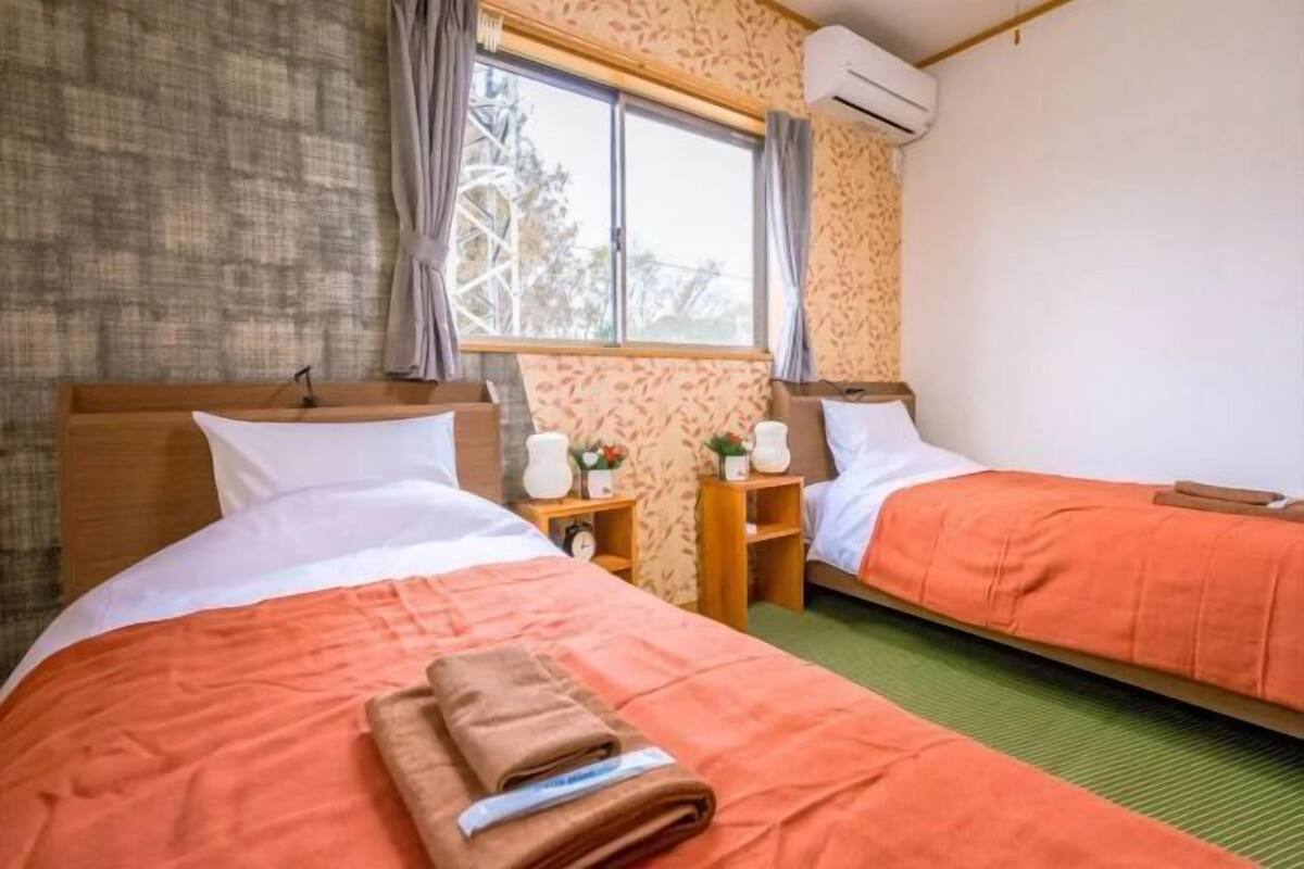 Quarto de hotel com duas camas, cada uma com colcha laranja, cabeceira de madeira e mesa de cabeceira com relógio e uma pequena planta.