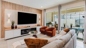 A modern living room with a large flat-screen TV, comfortable seating, and a view of the outdoor balcony.