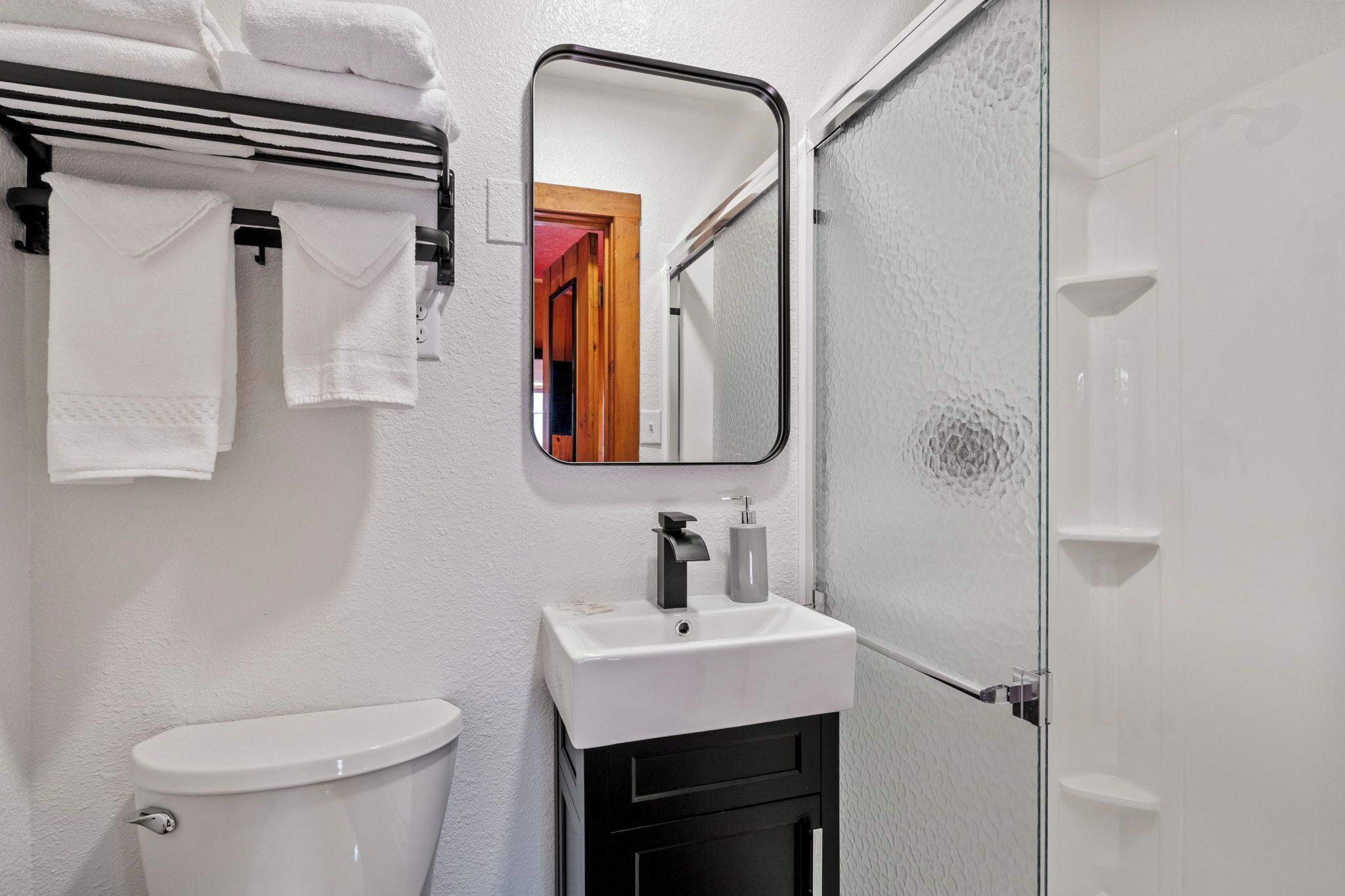 Een badkamer met een wit wastafel, een zwart kastje en een spiegel erboven. Handdoeken hangen aan een rek en er is een glazen douchedeur.
