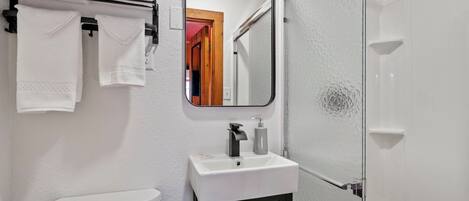 A bathroom with a white sink, black cabinet, and a mirror above it. There are towels hanging on a rack and a glass shower door.