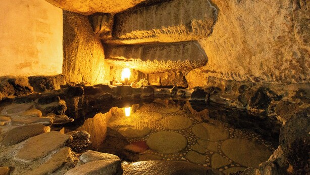 Eine unterirdische Kammer mit einer zentralen Lichtquelle, Steinwänden und einer spiegelnden Wasseroberfläche.