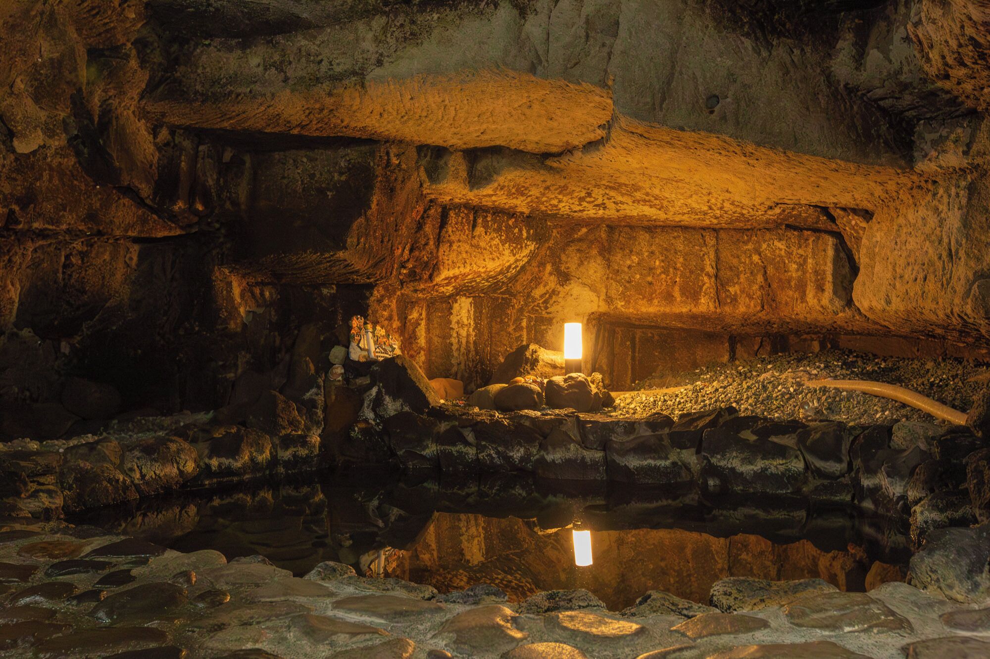 A cave interior with a central hut-like structure, a reflective water surface, and a cave wall.
