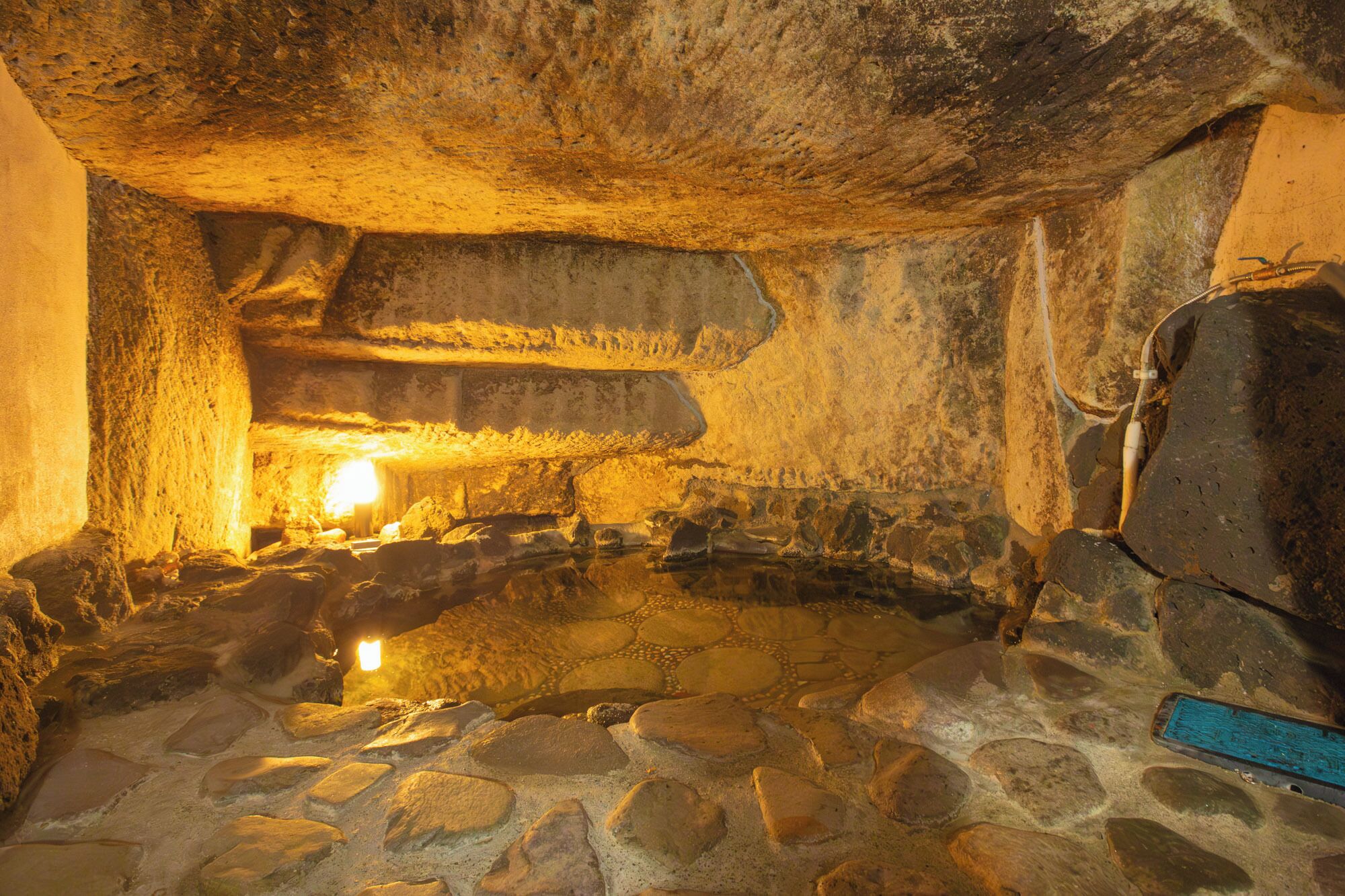 A stone-walled room with a central stone basin, possibly a hot spring, and a large, dark object on the right.