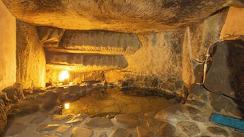 A stone-walled room with a central stone basin, possibly a hot spring, and a large, dark object on the right.