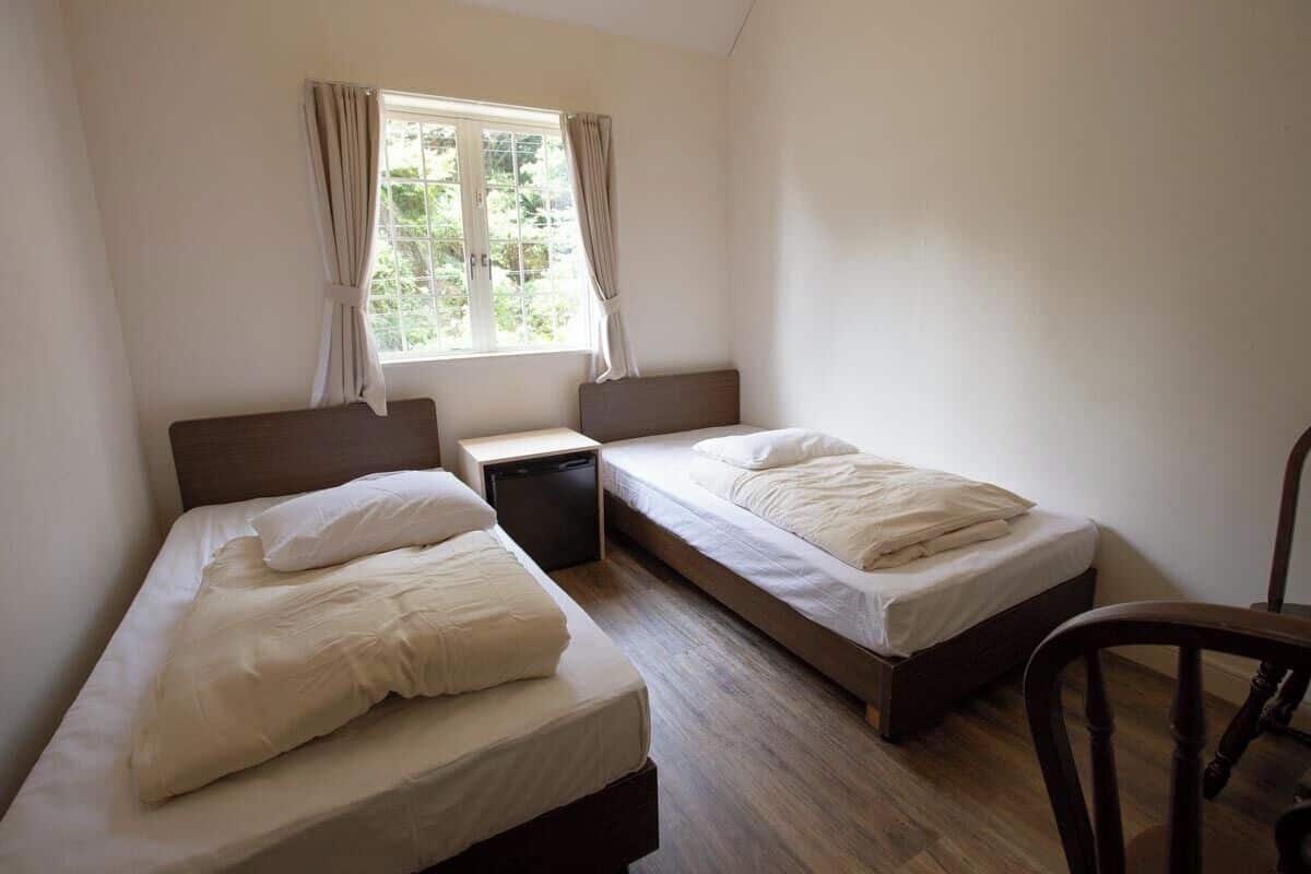 Una habitación con dos camas individuales, una mesita, una silla y un ventanal con cortinas.