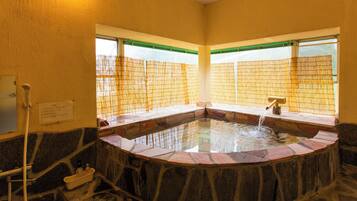 A stone hot tub with a tiled interior, two windows with green frames, and a wall-mounted faucet.